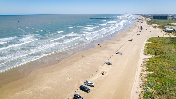Een strand met een lange zandvlakte, een steiger die in de oceaan reikt, en meerdere geparkeerde voertuigen langs de weg.