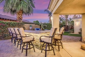 Une terrasse extérieure avec quatre chaises et un barbecue, entourée de palmiers et d’un mur de pierre.