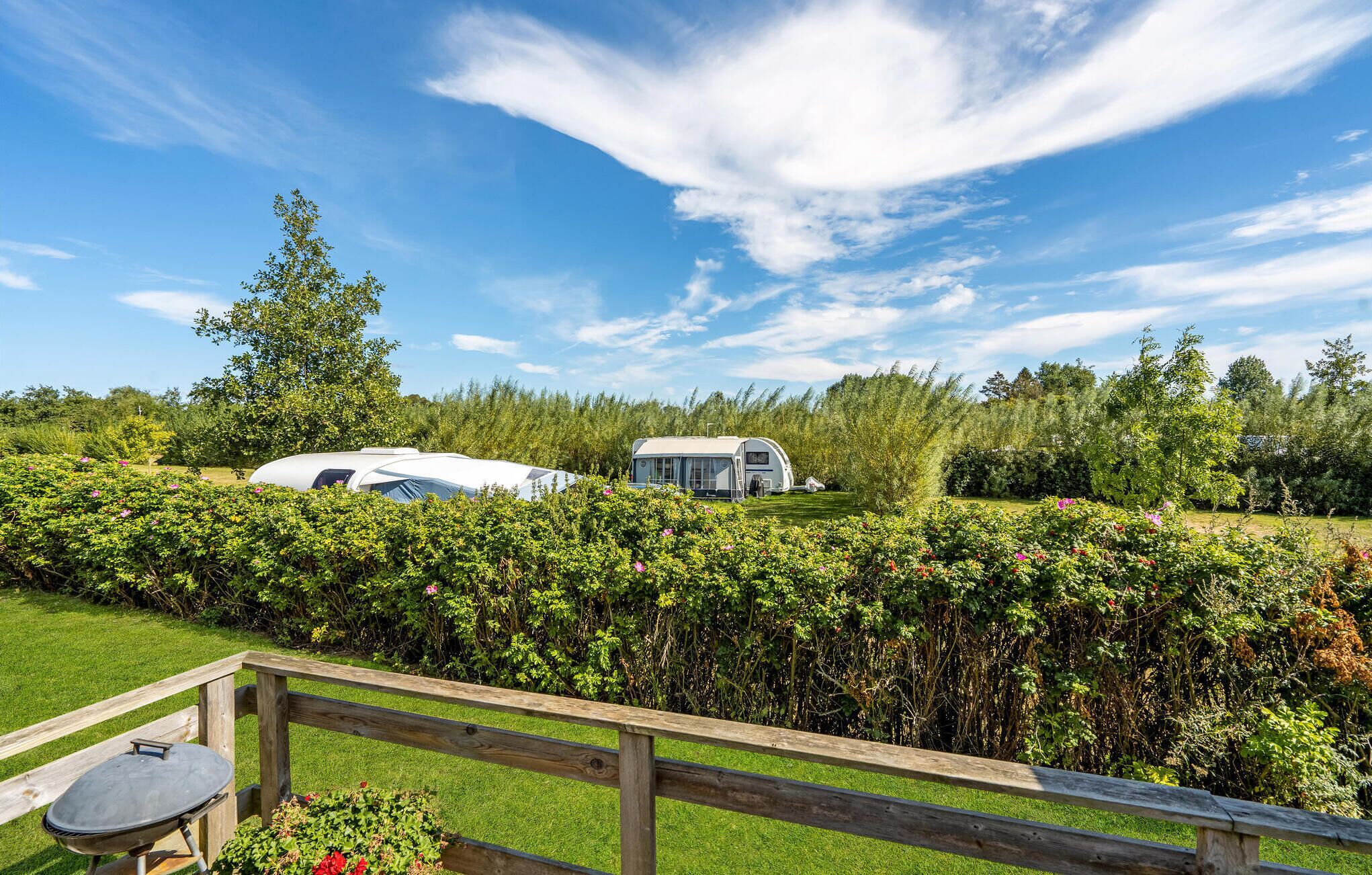 A campsite with a white RV and a smaller trailer, surrounded by lush greenery and a wooden fence.