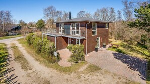 Une maison en bois à deux étages, dotée d’un balcon, entourée d’arbres et bordée d’un chemin de gravier.