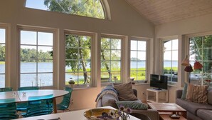 A spacious living room with large windows overlooking a lake, a dining area with a glass table, and a cozy seating area with a TV.