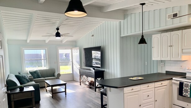 A modern kitchen with white cabinetry, a black countertop, and a built-in oven. There is a dining area with a table and chairs, and a living room with a sofa and a TV.