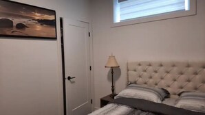 A bedroom with a bed, a nightstand, a lamp, a painting, and a window. - Adorable 1 bed room legal basement suite (Saskatoon)