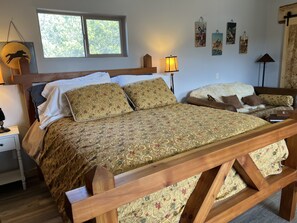 Ein hölzernes Bett mit einem gemusterten Überwurf, ein Nachttisch mit einer Lampe und ein Fenster mit Blick auf Bäume.