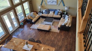 A spacious living room with a large wooden dining table, multiple sofas, and a view of the outdoors through large windows. - Secluded Lakefront Retreat on 60 Acres Private Dock, Fishing & Wildlife (Gravois Mills)