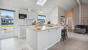 Une cuisine moderne dotée d’un îlot central, de meubles blancs et offrant une vue sur une maison à travers une porte vitrée.