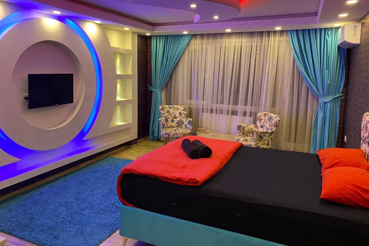 A modern hotel room with a bed, two armchairs, a flat-screen TV, and a large window with curtains.