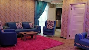 Ein Wohnzimmer mit zwei Sofas, einem Couchtisch und einem roten Teppich. Die Wände sind mit ornamentalem Tapetenmuster versehen und der Boden ist aus Holz.