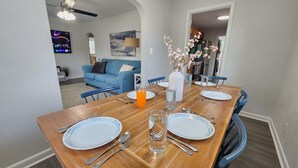 Un coin repas avec une table en bois mise pour quatre personnes, comprenant des assiettes, des couverts et des verres. Un vase rempli de fleurs est posé sur la table, et un ventilateur de plafond est installé au-dessus.