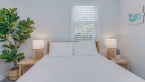 Une chambre à coucher comprenant un lit, deux tables de chevet, une plante en pot, une fenêtre avec des stores et une œuvre d’art accrochée au mur.
