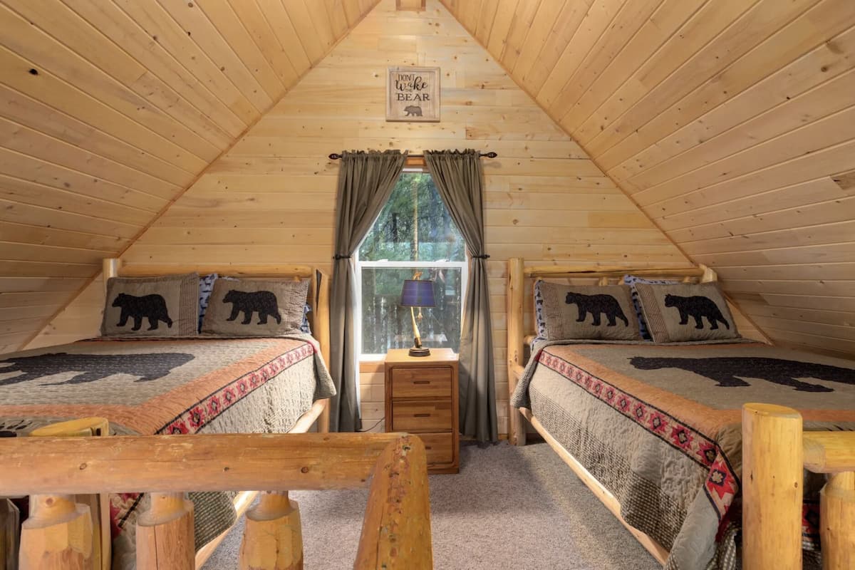 A cozy cabin room with two beds, each with a bear-themed pillow, a small wooden nightstand, and a window with curtains.