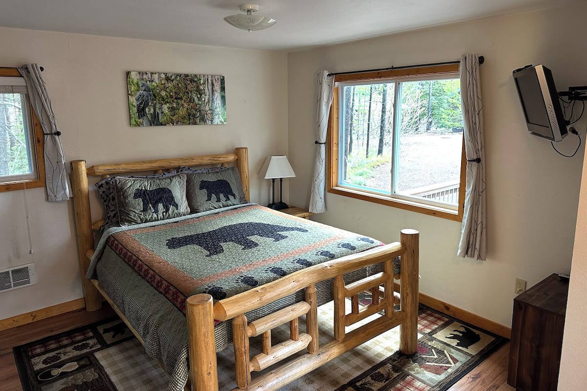 A bedroom with a wooden bed, a window with curtains, a painting on the wall, and a television mounted on the wall.