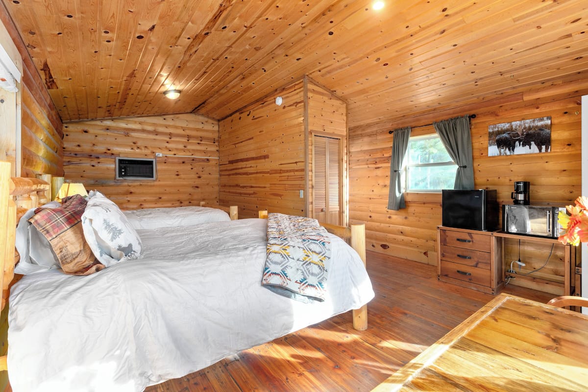 A wooden cabin room with a bed, a television, a coffee maker, and a window with curtains.