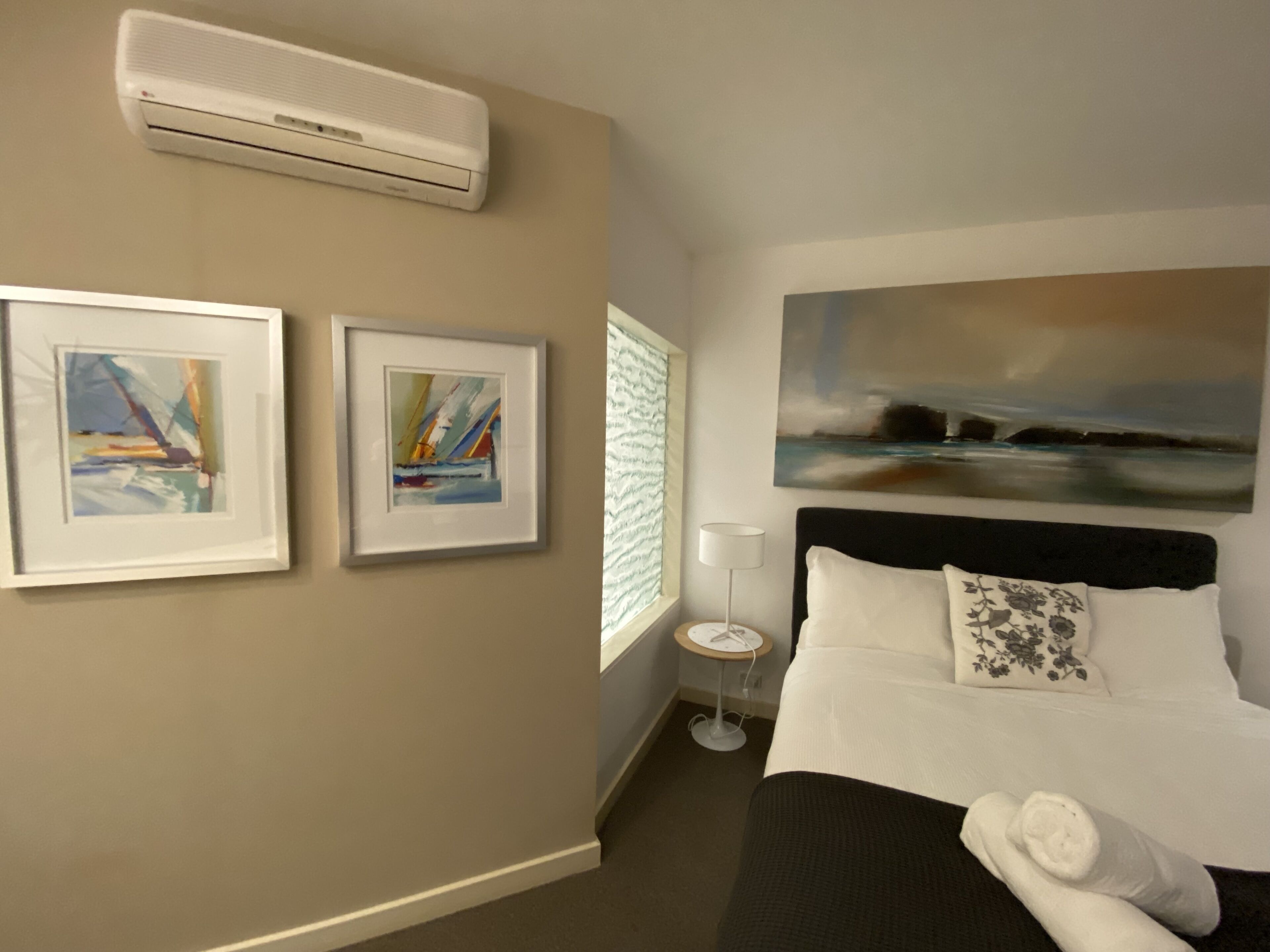 Ein Hotelzimmer mit Bett, zwei gerahmten Bildern, einem Fenster mit Jalousien, einem Nachttisch mit Lampe und einer Klimaanlage.