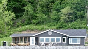 A cabin with a stone wall, a covered patio with outdoor furniture, and a view of a lake. - Reel Paradise at Shore Slough (Lansing)