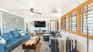 A living room with a blue sofa, a wooden coffee table, and a grey couch. There is a TV on the wall and a window with blinds. - Desert Ruby: Stargaze, Spacious, Hot Tub, Fire pit, Fenced, Pet Friendly (Yucca Valley)