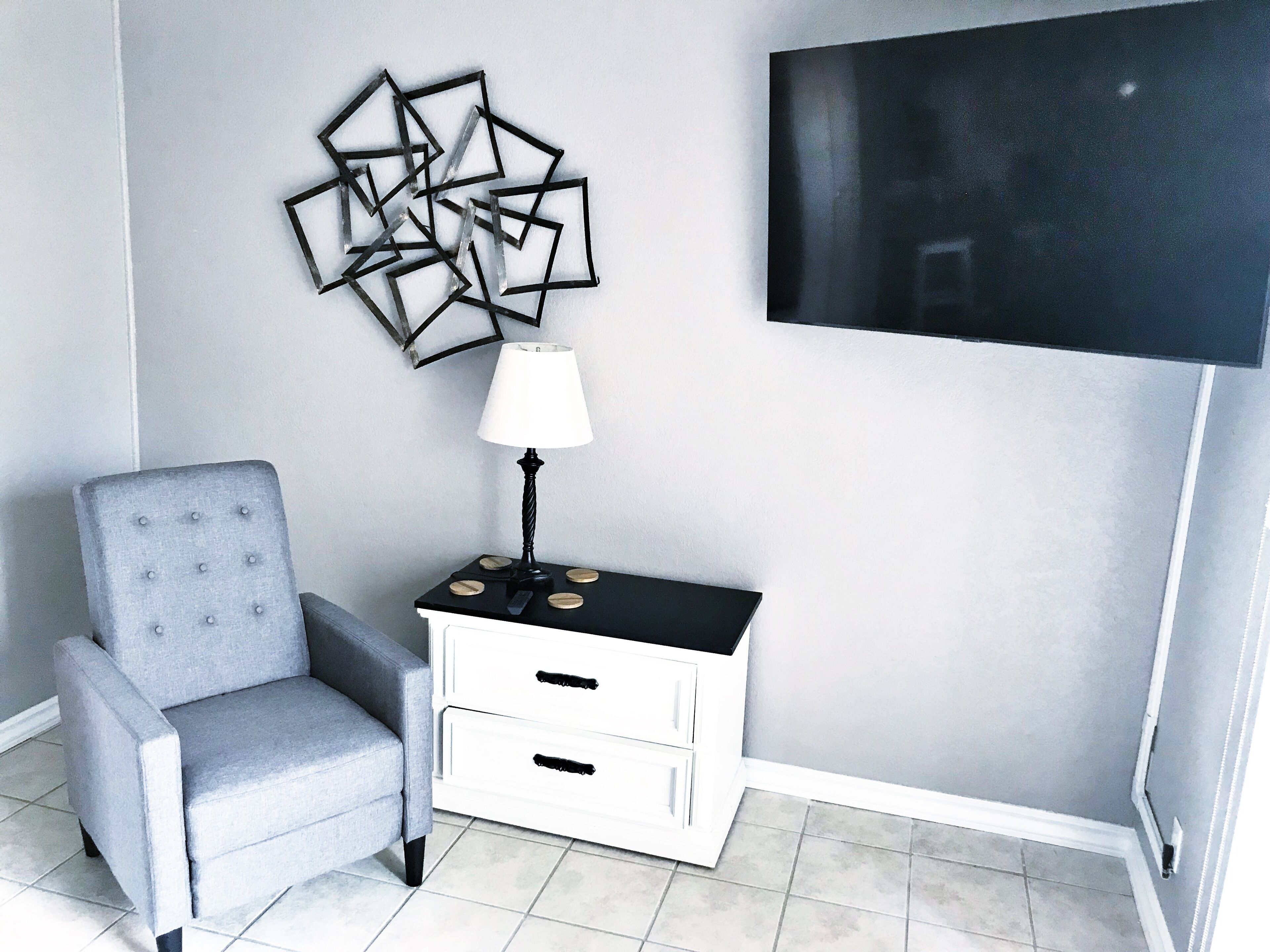 A modern living room with a grey armchair, a black and white side table with a lamp, and a wall-mounted TV.