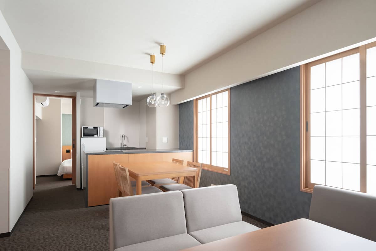 A modern hotel room with a dining area, kitchenette, and a large window providing natural light.