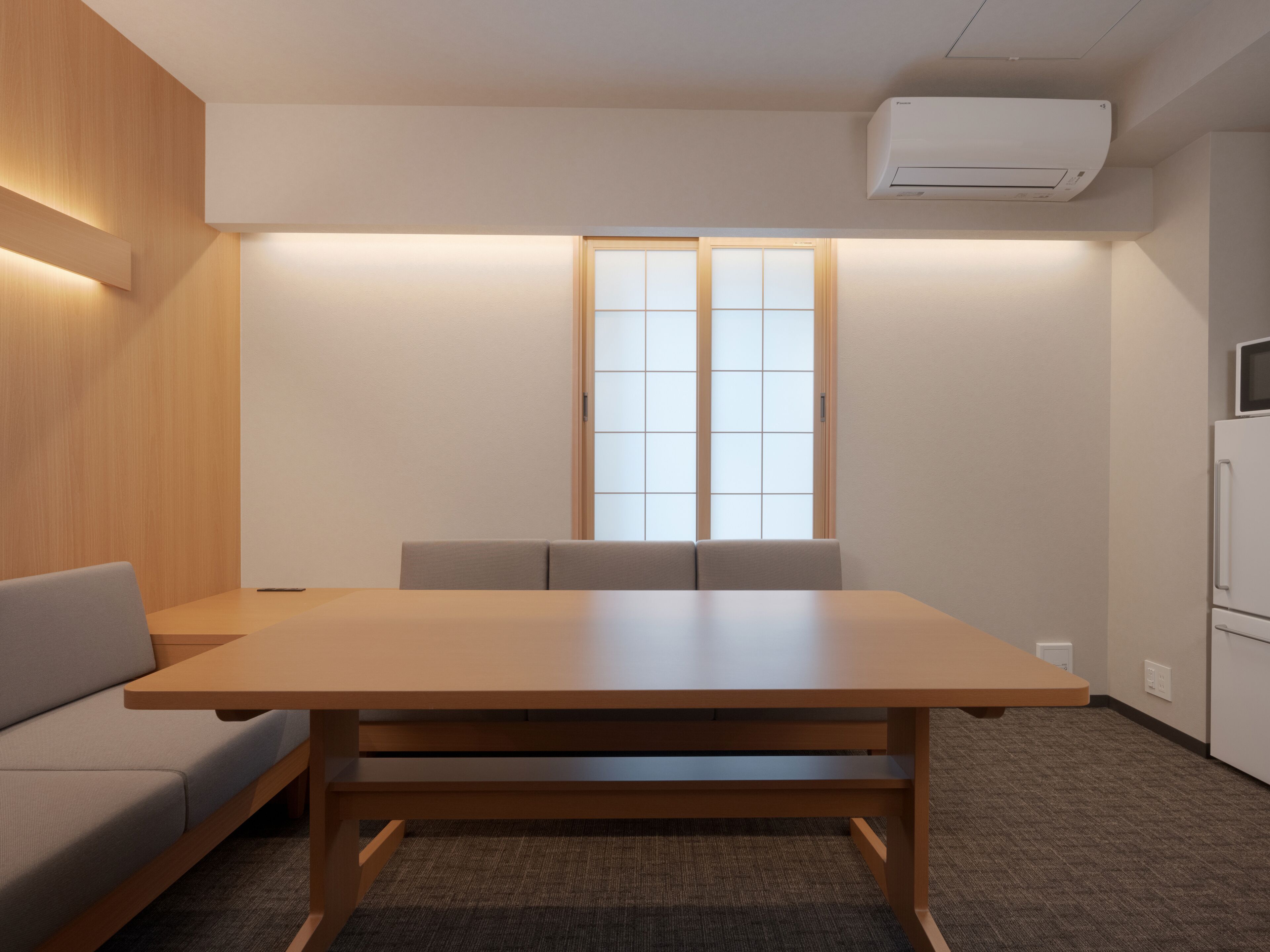 A modern meeting room with a wooden table, cushioned chairs, a sofa, a large window with frosted glass, and an air conditioning unit.