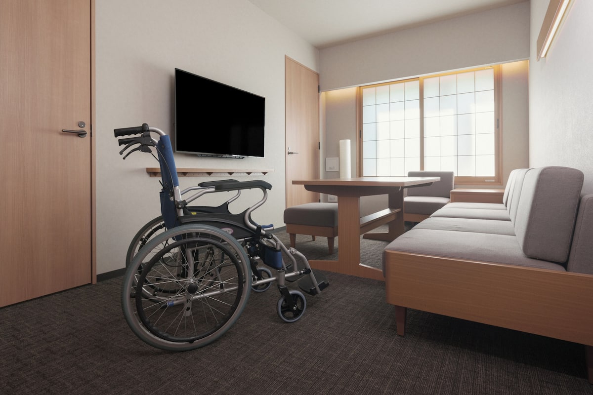 A wheelchair-accessible room with a TV, a sofa, a table, and a window with blinds.
