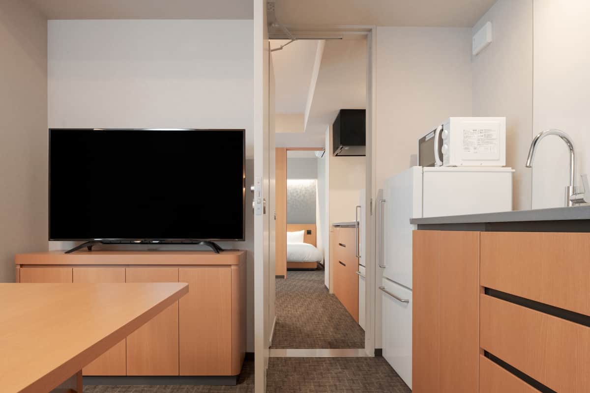 A compact hotel room with a flat-screen TV, a kitchenette with a fridge and microwave, a small dining table, and a carpeted floor.
