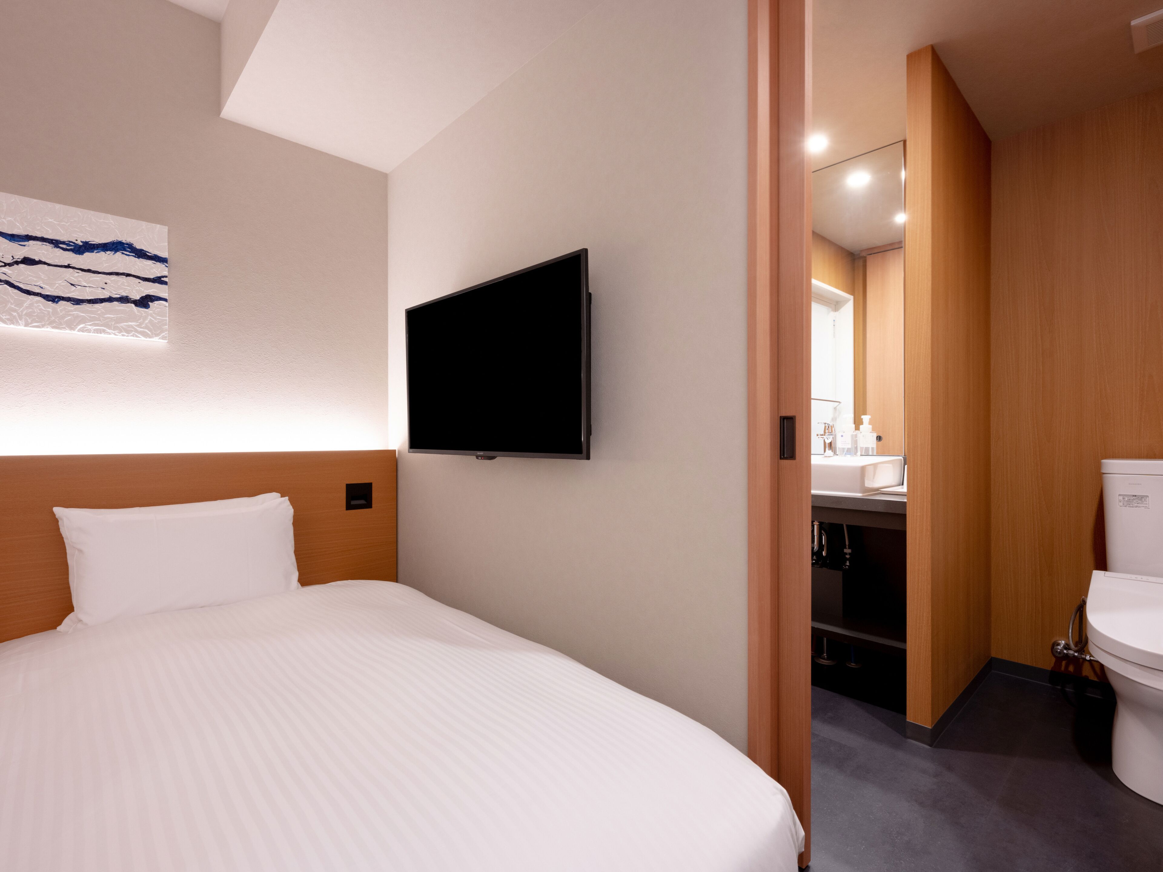 Ein Hotelzimmer mit Bett, einem an der Wand montierten Flachbildfernseher und einem durchgehenden Türspalte sichtbaren Badezimmer.