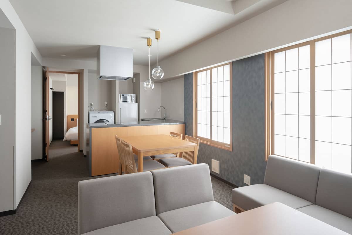A modern hotel room with a dining area, kitchenette, and a large window providing natural light.