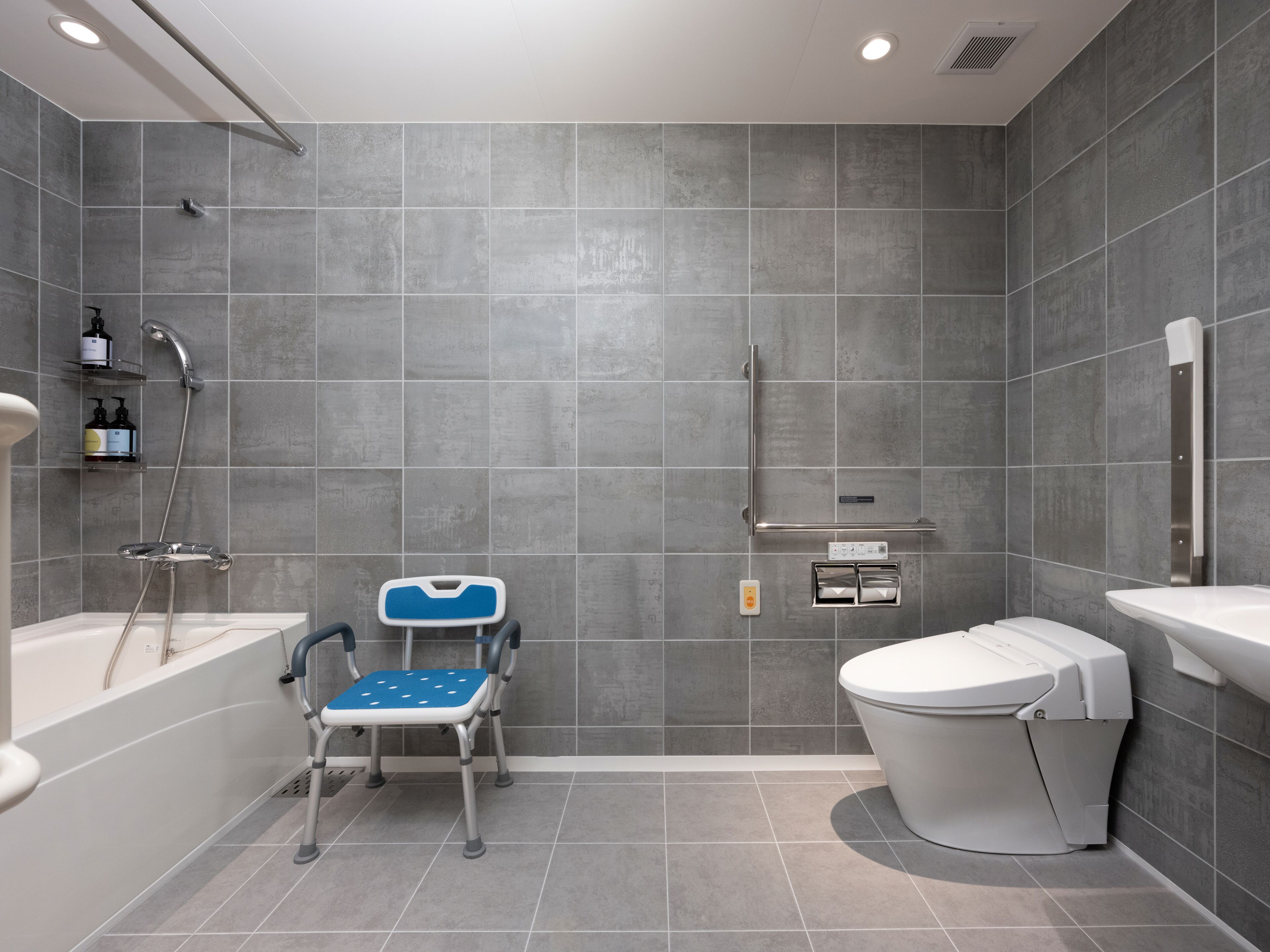 A modern bathroom with grey tiles, a white bathtub, a white toilet, and a blue mobility aid.