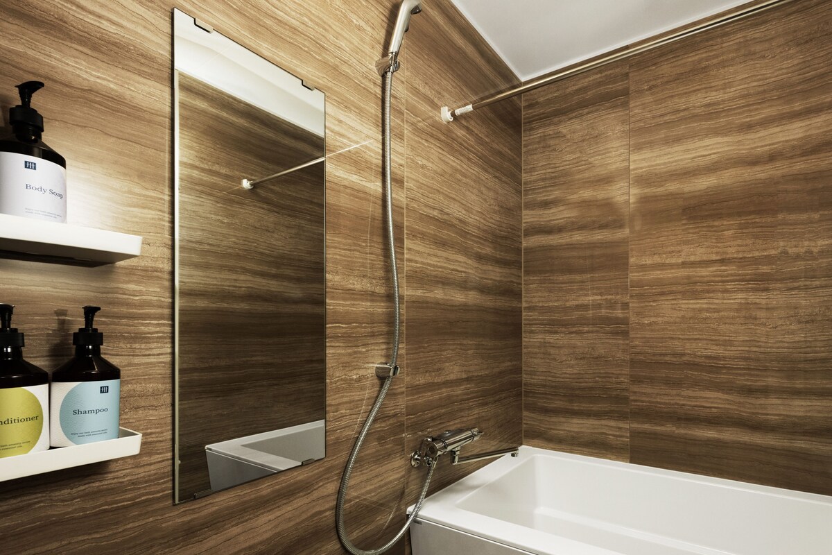 Un baño moderno con pared de madera, una bañera blanca y ducha con manguera. Hay estantes con botellas de jabón de baño y champú.