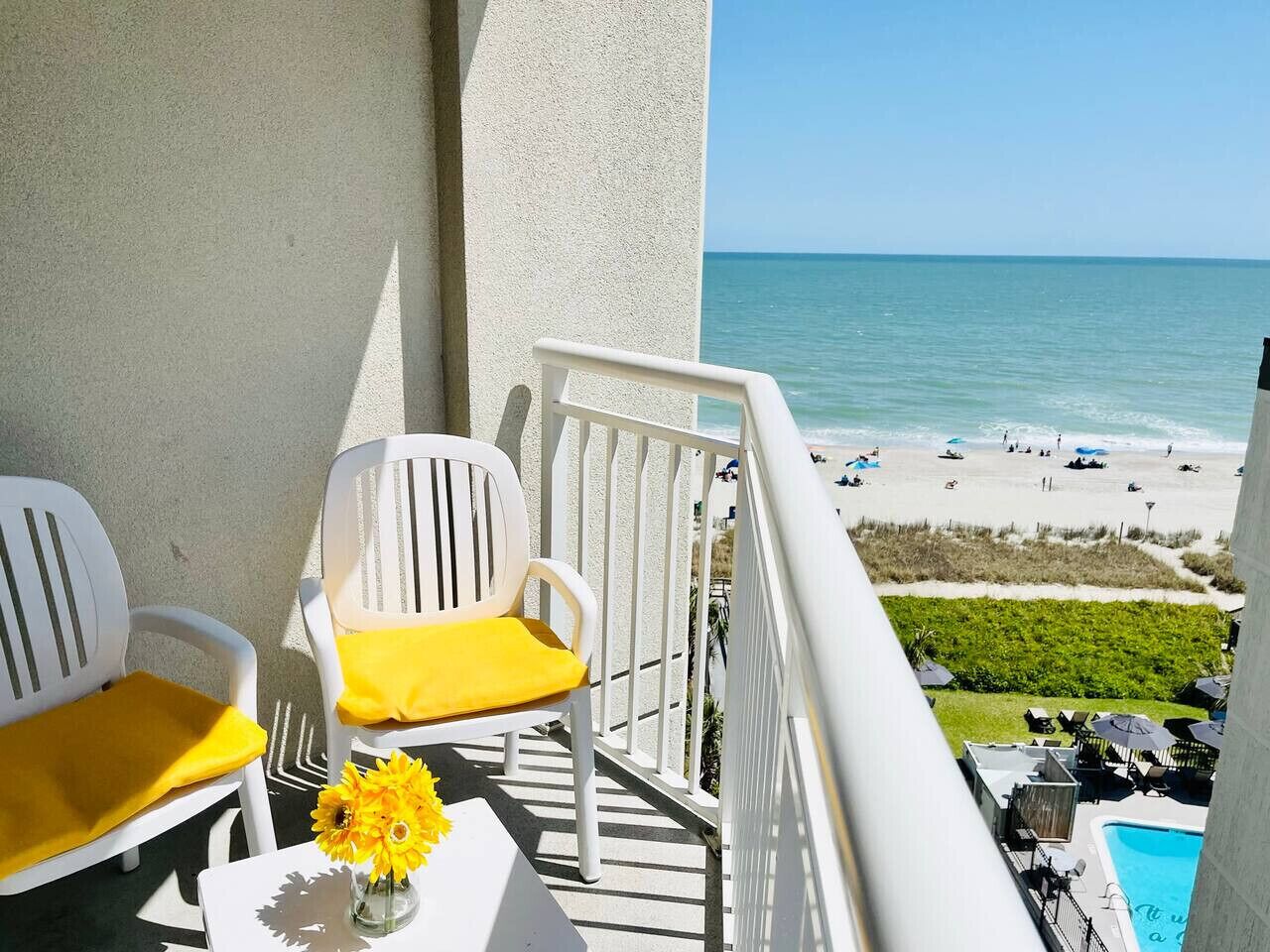 A balcony with two white chairs, one with a yellow cushion, and a vase with yellow flowers. The balcony overlooks a beach with people and umbrellas.