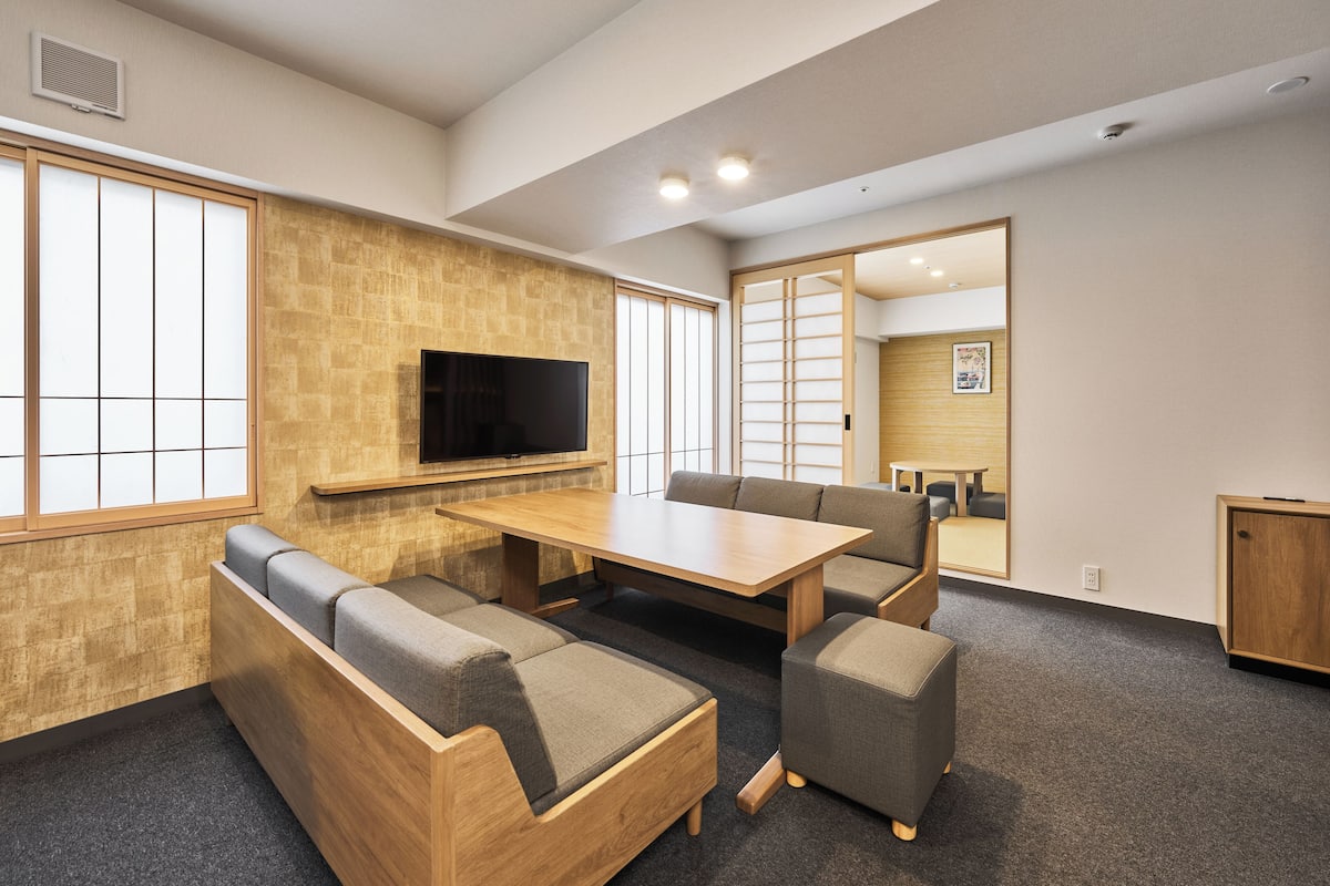 A modern interior with a wooden table and cushioned seating, a flat-screen TV mounted on the wall, and large windows providing natural light.