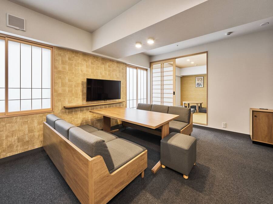 A modern interior with a wooden table and cushioned seating, a flat-screen TV mounted on the wall, and large windows providing natural light.