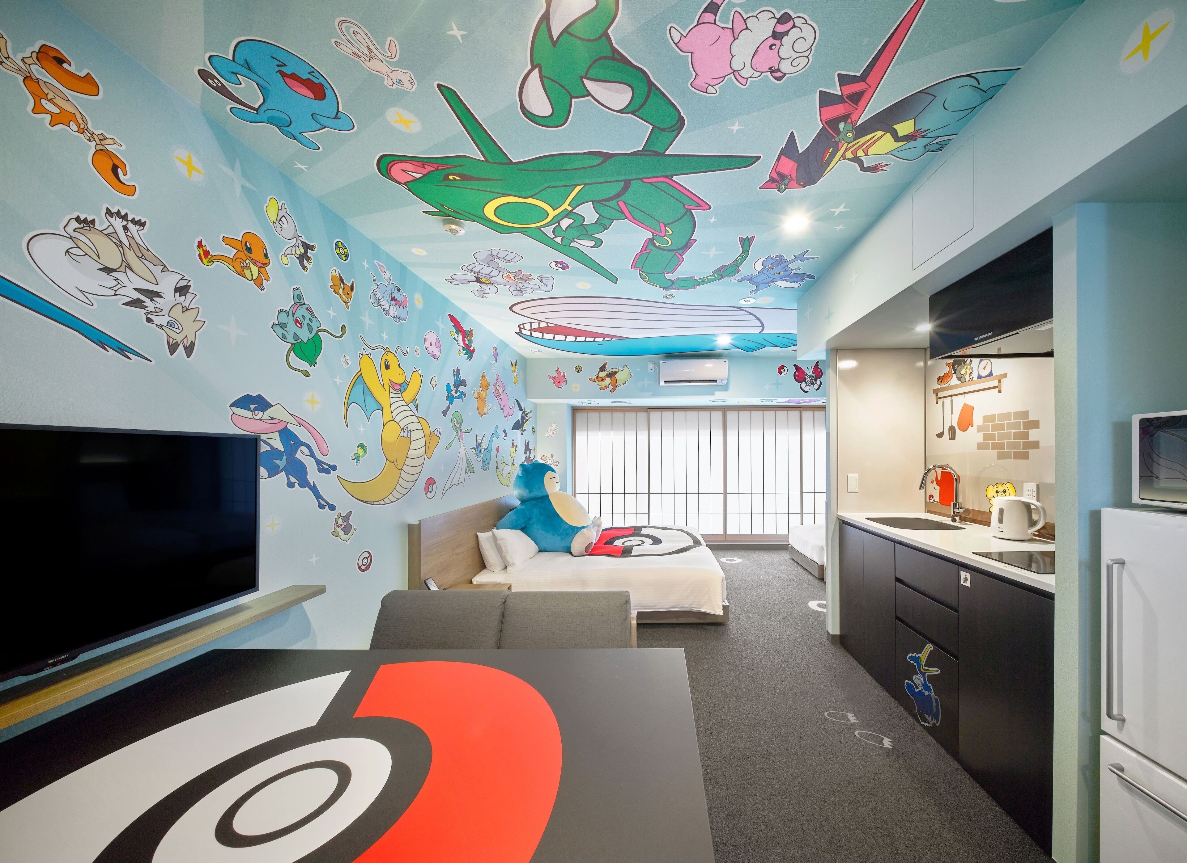 A room with a large TV, a sofa, a dining area with a kitchen, and a ceiling with colorful wall art.
