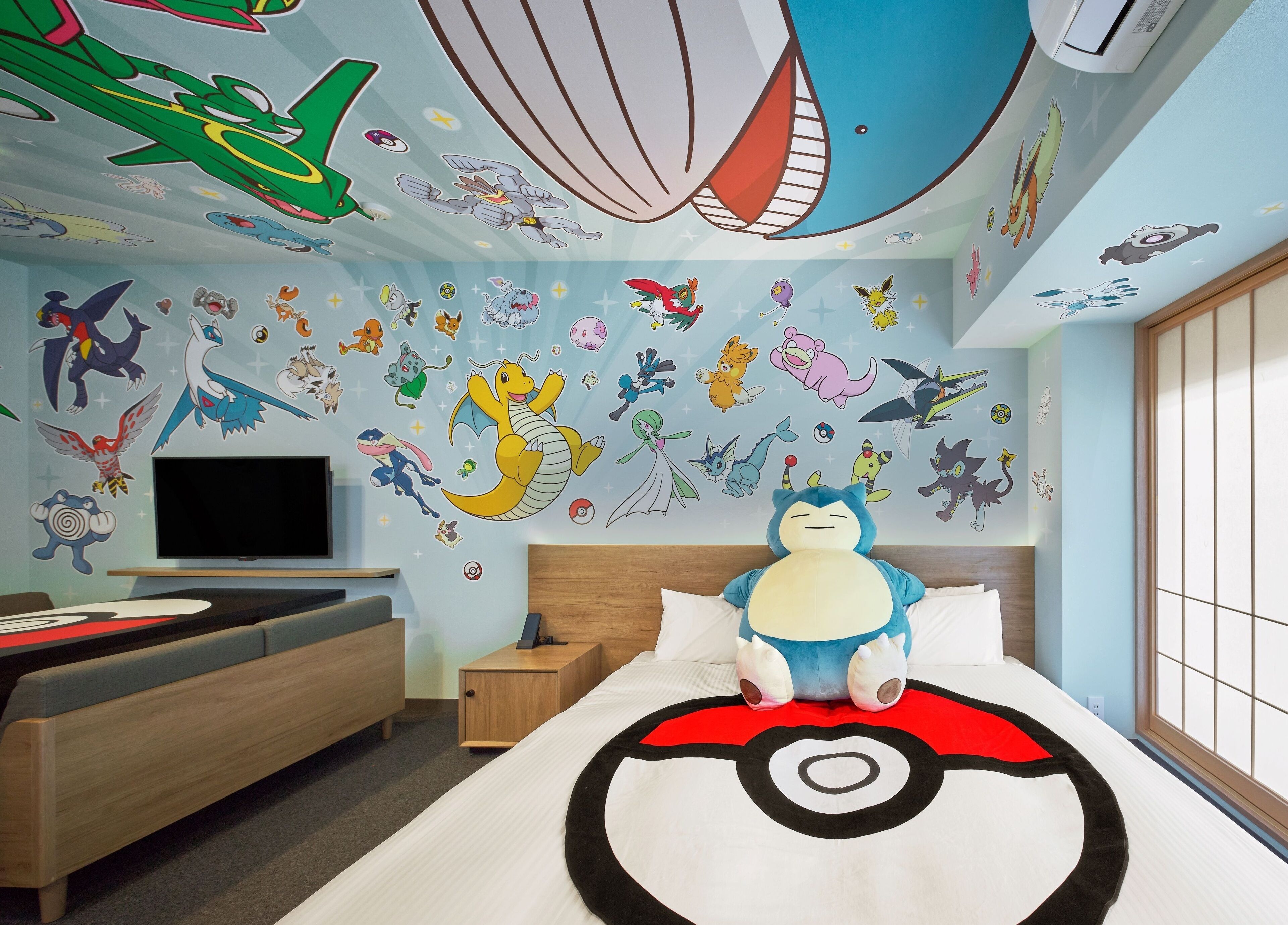 Un dormitorio con una cama, almohada temática de Pokemon, televisión, mesita de noche de madera y ventana con vistas.