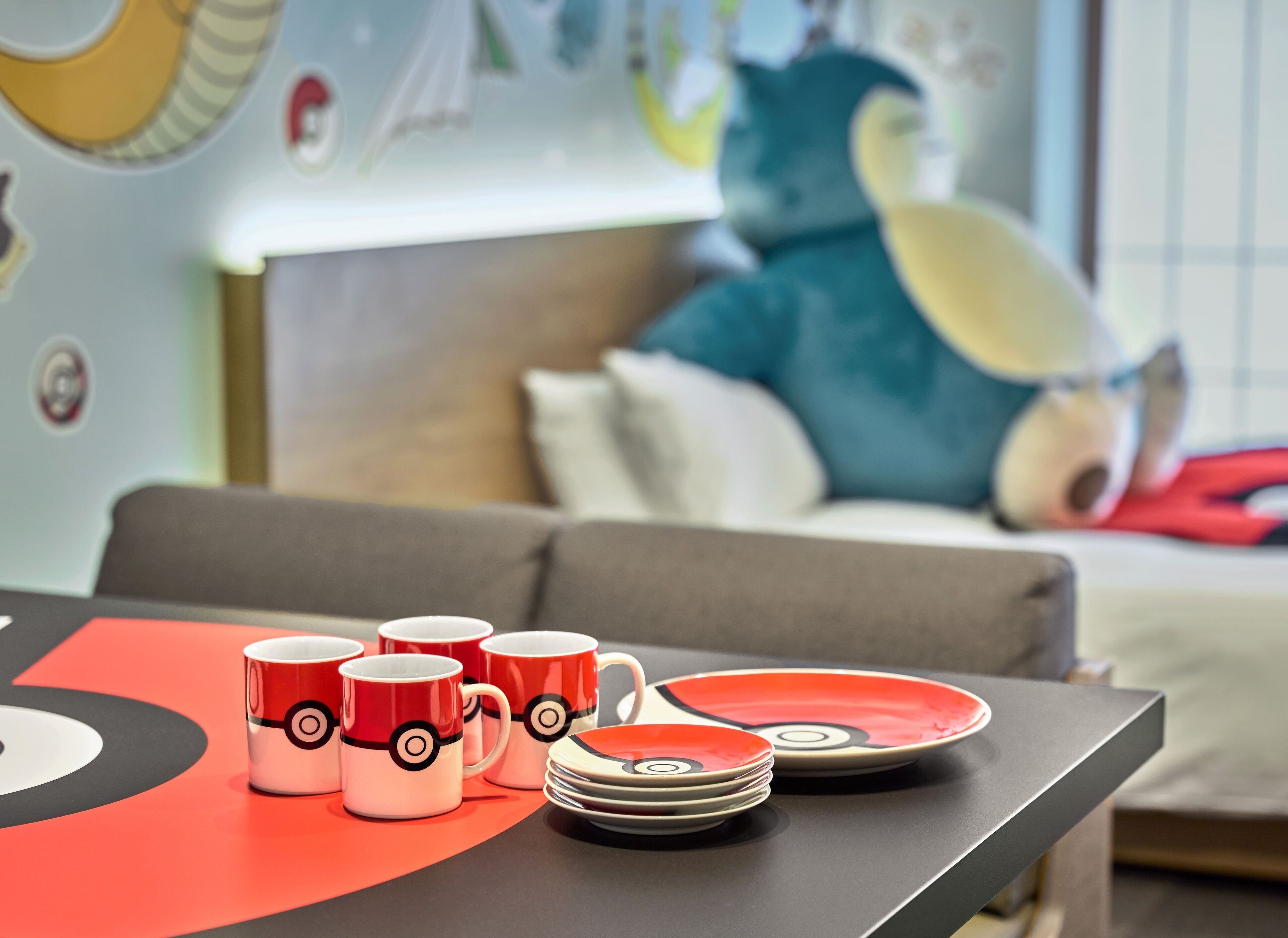 A table with red and white mugs and plates with a circular design, a grey sofa, and a large plush toy in the background.