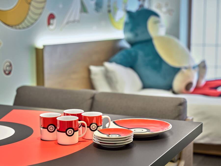 A table with red and white mugs and plates with a circular design, a grey sofa, and a large plush toy in the background.