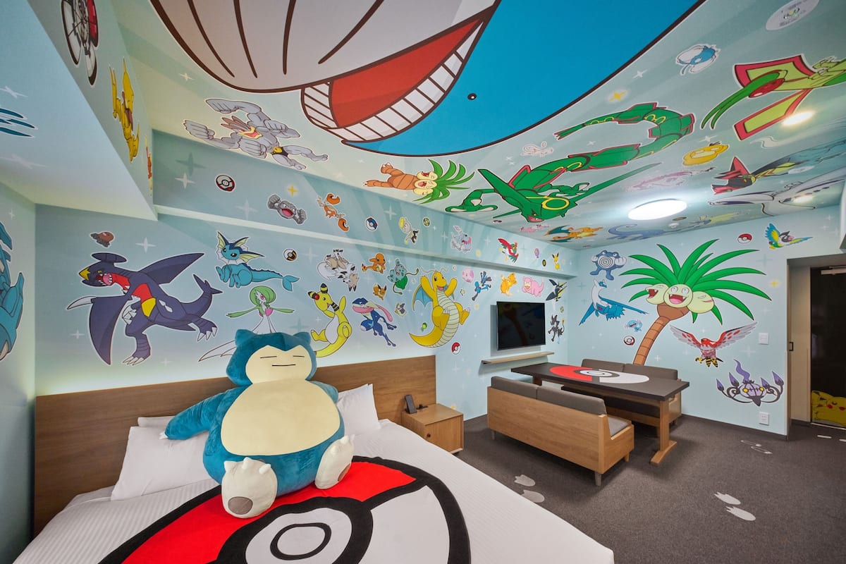 A room with a bed featuring a Pokemon-themed pillow, a TV, and a wall decorated with various Pokemon characters.