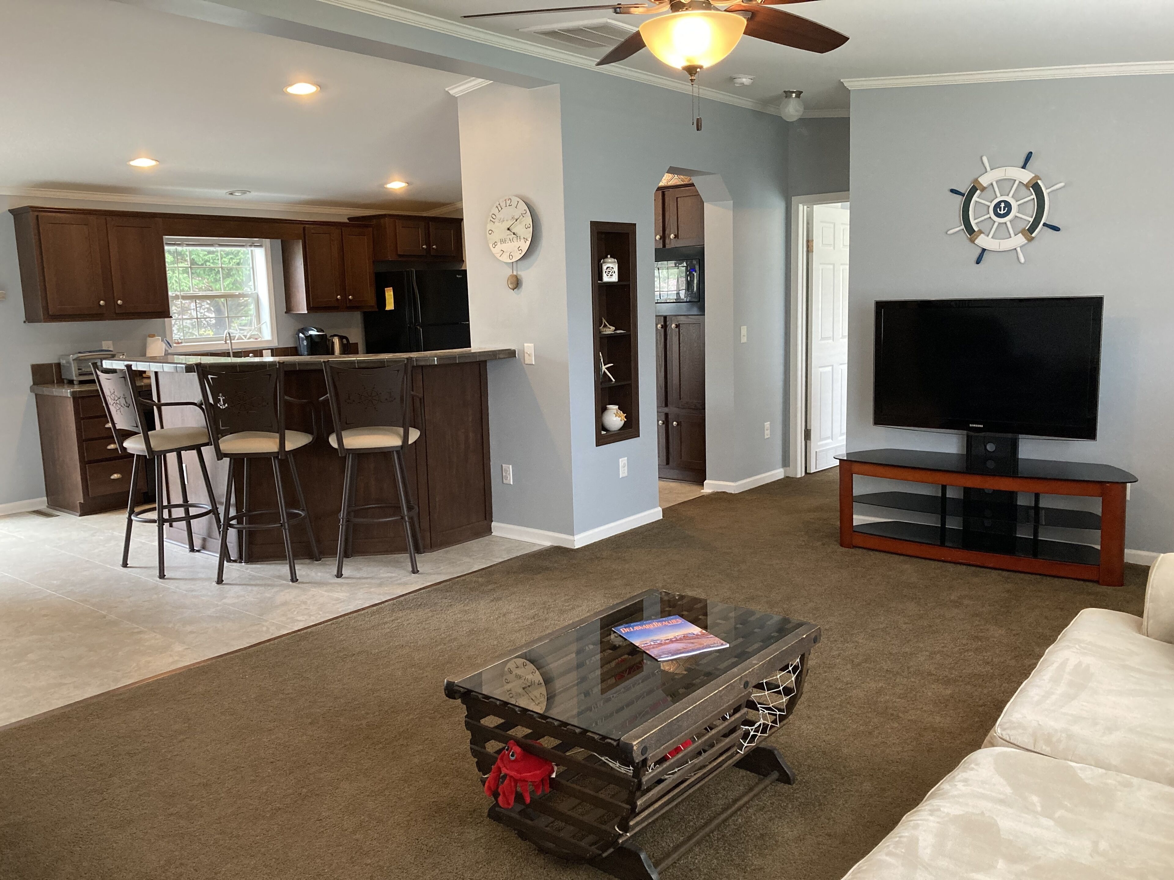 A modern living room with a glass coffee table, a television on a wooden stand, and a kitchen area with dark wooden cabinets and a bar stool.