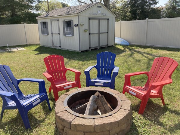 Ein Garten mit Feuerstelle, vier Adirondack-Stühlen und einem Schuppen mit der Aufschrift 'Bethany Beach'.