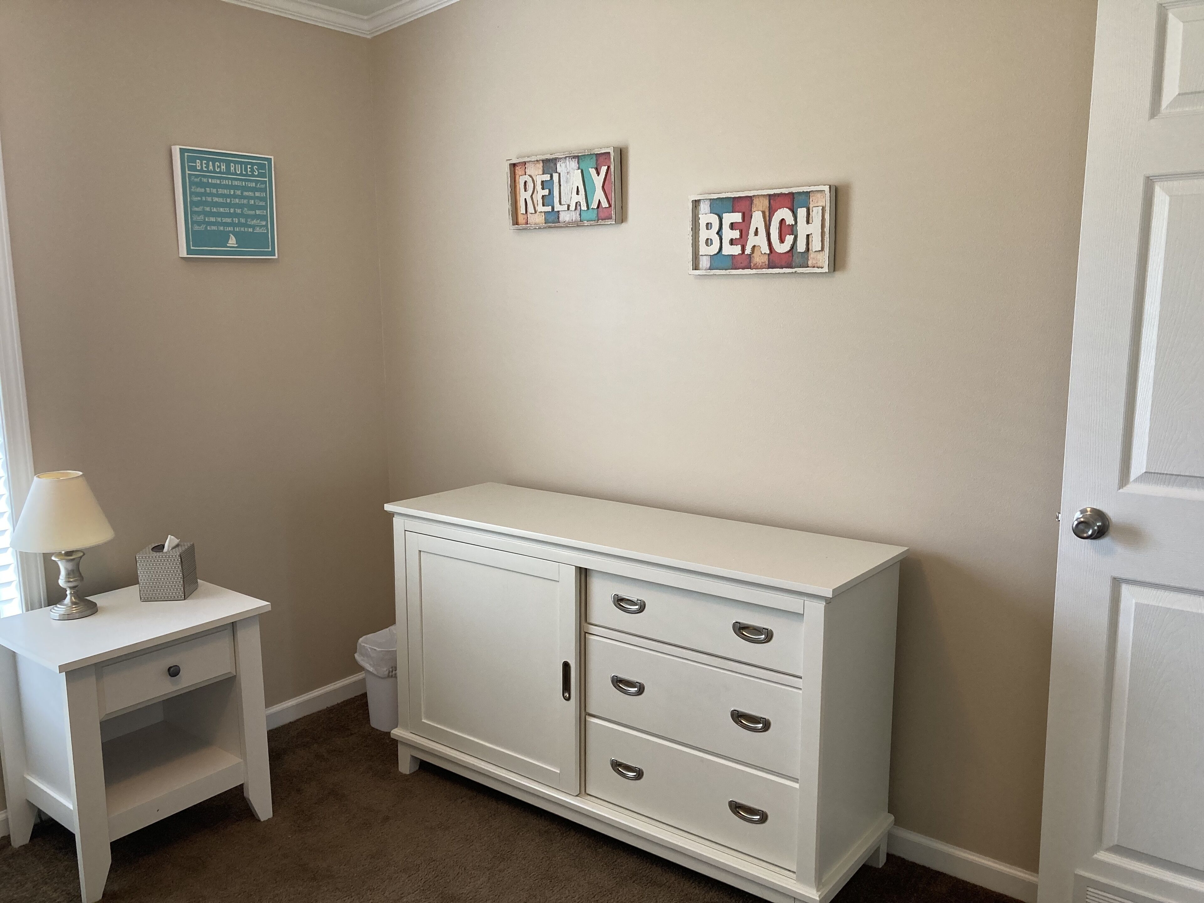 Een kamer met een witte dressoir, een klein bureau met een lamp, en wanddecoraties met de tekst 'BEACH RULES', 'RELAX' en 'BEACH'.