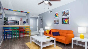 A modern living room with a white coffee table, an orange sofa, and a kitchen area with colorful chairs.