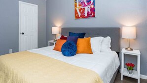 A neatly made bed with a white comforter and several colorful pillows, a white bedside table with a lamp, and a wall art piece above the bed.