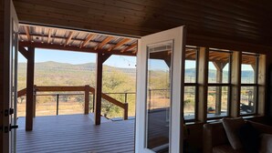 Terraza de madera con vistas a un paisaje montañoso, puerta corrediza de vidrio y sofá de cuero con cojines.