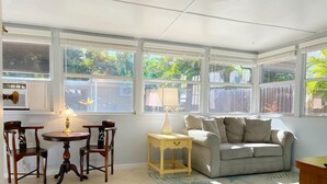 A sunlit room with a grey sofa, wooden chairs, a small table with a lamp, and a view of greenery outside. - Sarasota Gateway Beach House (Sarasota)