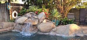 A rock waterfall feature in a garden with a wooden fence, potted plants, and outdoor furniture.