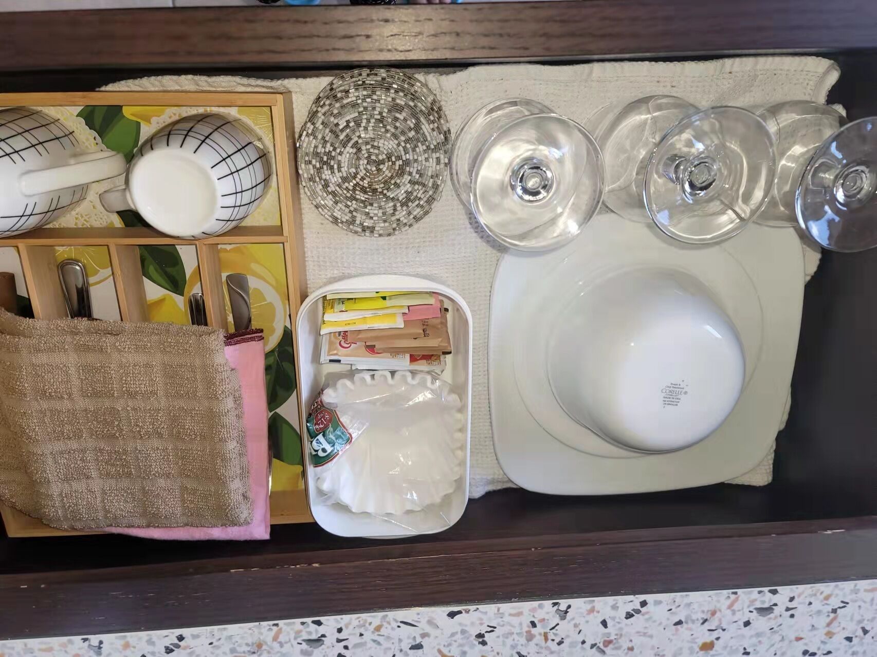 Keittiön työtasolla on erilaisia esineitä, kuten kudottu alusta, astiateline, valkoinen kulho, pieni lautanen lusikan kanssa, kangaskassi ja joitain pakattuja elintarvikkeita.