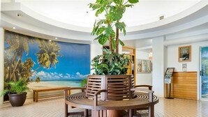 Un vestíbulo con una mesa redonda de madera, dos sillas, una planta grande en maceta, un banco y un mural con una escena de playa.