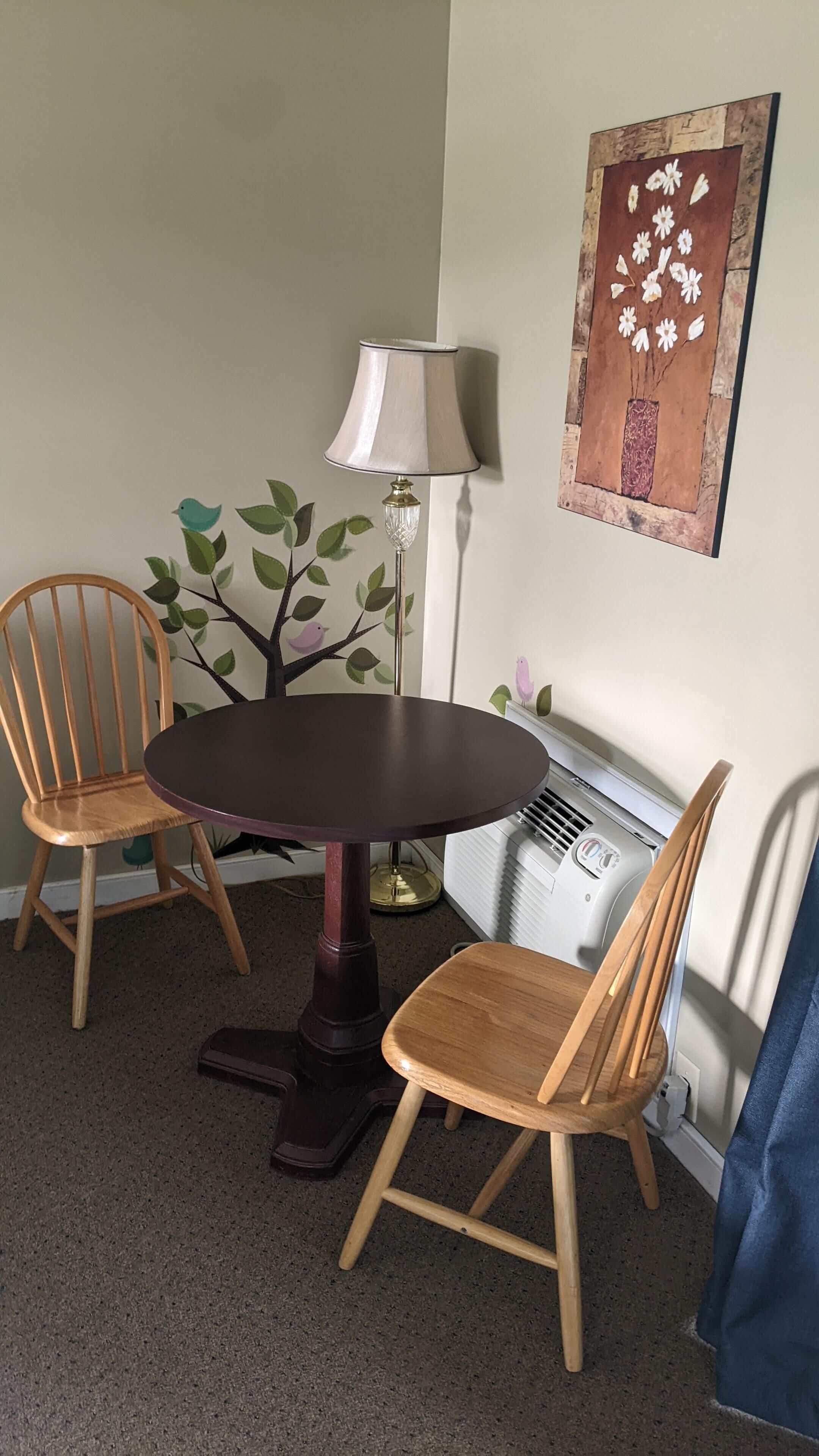 Un coin repas comprenant une table ronde, deux chaises en bois, un lampadaire et une œuvre d’art accrochée au mur.
