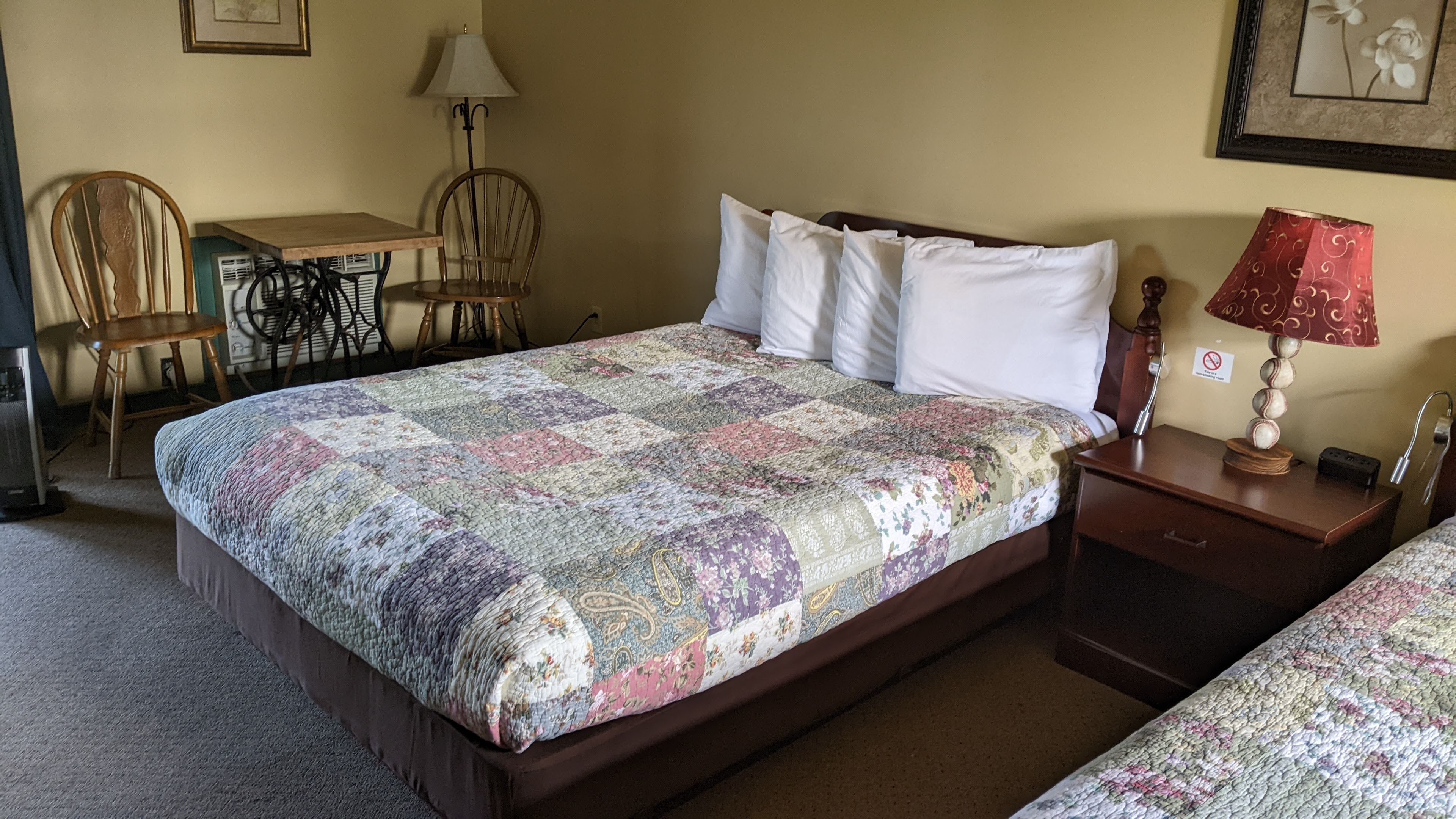 A neatly made bed with a floral quilt, a wooden nightstand with a lamp, and a chair with a table in the background.