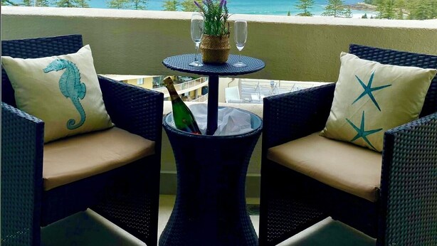 Een balkon met rieten meubels, een tafel met fles en glazen, en uitzicht op zee en gebouwen.